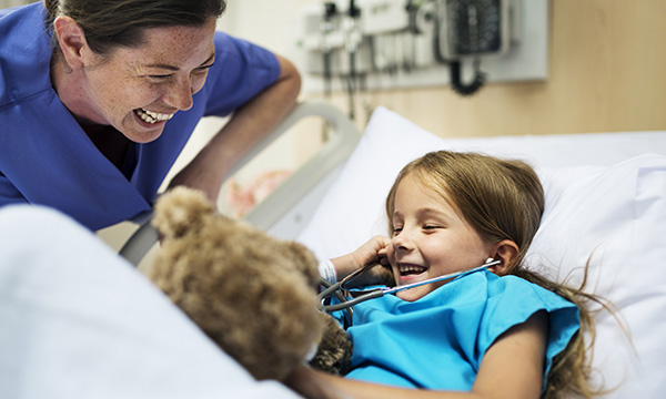 A laughing children’s nurse looks on as a smiling young girl listens to her teddy bear’s heartbeat with a stethoscope