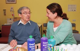 A nurse sits talking to a nursing home resident as he undertakes a craft activity