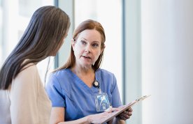 A patient, seen from behind, talks in confidence to a nurse in uniform who is holding a clipboard