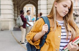 A female student looks downcast and worried as she walks alone outside of a university building while other students chat in the background, suggesting a nursing student who is isolated or struggling with their mental well-being