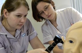 A nursing student holds an otoscope to the ear of a manikin as her trainer looks on