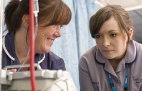 A nursing student looks at an IV monitor as she listens to a senior nurse while learning on placement