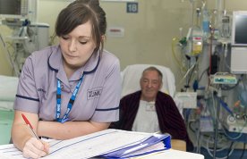 Record keeping in nursing: a nursing student leans on a table writing in a patient’s notes as the patient lies in a hospital ward bed behind her