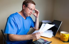 Nurses’ pay and pension worries: concerned nurse sits at desk, head in hand as he read documents that could be bills or bank statements