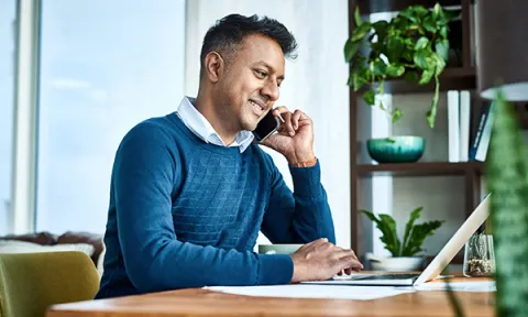A man sitting in a home office talking on a phone, while looking at patient notes, as with an occupational health nurse working from home