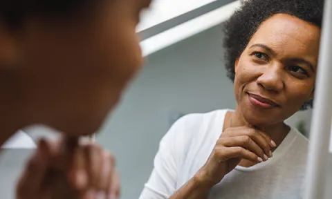 A smiling woman appears deep in thought as she looks at her reflection in a mirror