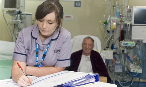 Record keeping in nursing: a nursing student leans on a table writing in a patient’s notes as the patient lies in a hospital ward bed behind her