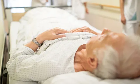 An older person lies on a bed in an emergency department corridor because there is nowhere else to put them. This is part of the emergency department crisis affecting patients and nurses