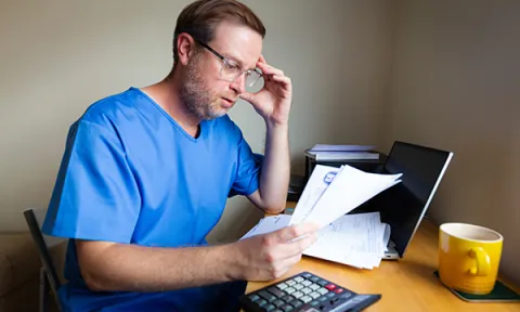 Nurses’ pay and pension worries: concerned nurse sits at desk, head in hand as he read documents that could be bills or bank statements