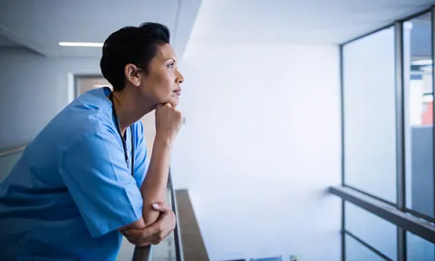 NHS nurse redundancy: pensive nurse leans on a corridor railing as she gazes out of a window