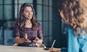 A nurse sitting at a desk in a job interview gestures while speaking to an interviewer