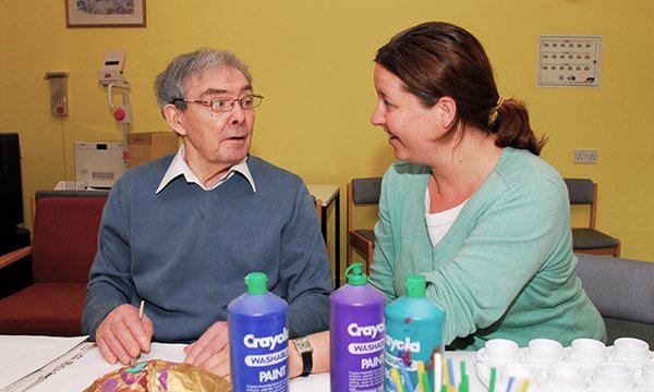 A nurse sits talking to a nursing home resident as he undertakes a craft activity
