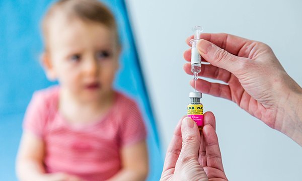 hands of a healthcare professional in foreground preparing to administer MMR vaccine to baby out of focus in the background