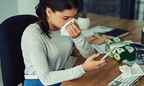 woman sits at her desk, looking at her phone and blowing her nose