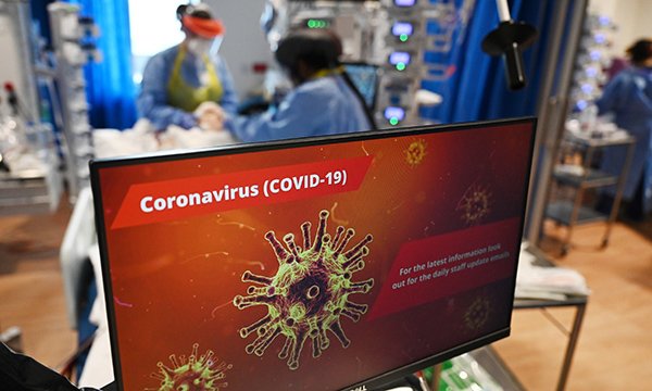 Picture of COVID-19 notice on a glass door. In the background clinical staff wearing personal protective equipment look after a patient in an intensive care unit.