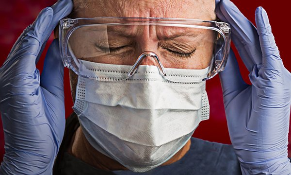 Nurse wearing face mask and goggles holds hands to temples and appears distressed