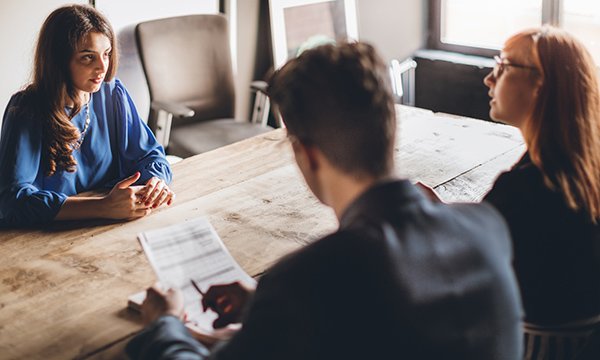 Picture shows a young woman sat at a table and being questioned