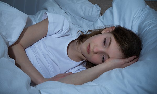 Picture sows a young girl with her head on a pillow, touching her head with her hand and looking sad 