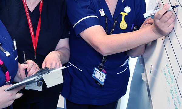 Picture sows three nurses gathered in front of a work roster, with one writing on the roster and another looking at a clipboard