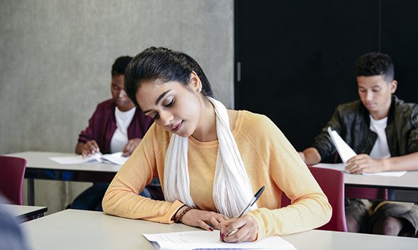 A young woman sits at a desk in a test centre, with two others at desks behind her, taking a language test for internationally trained nurses