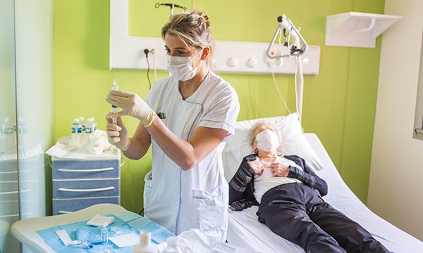 A nurse wearing gloves and a mask prepares a chemotherapy drug while standing at a table next to a hospital bed on which a male patient is lying