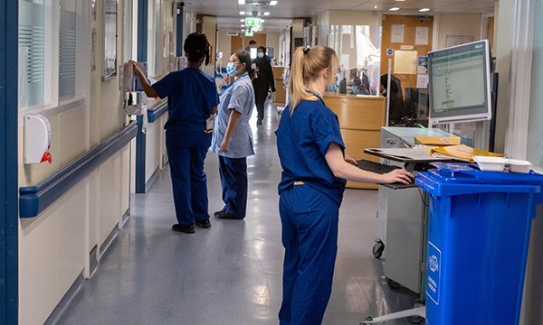Clinicians chat in a hospital corridor while a colleague nearer the camera calls up information on a computer screen