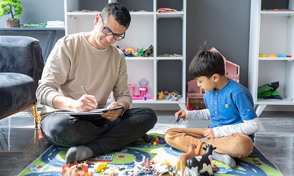A play therapist sits cross legged on the floor making notes as a young boy plays with some plastic animals