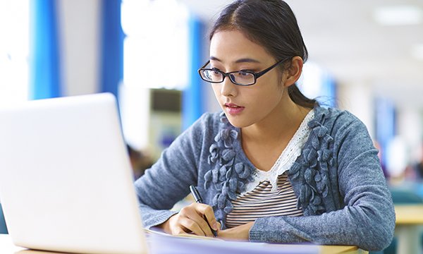 A student looks at an open laptop with pen in her hand to take notes