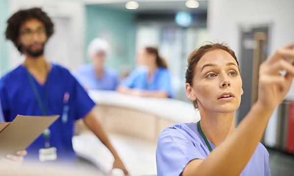 A nurse writes on the roster board with a blurred and busy ward behind her
