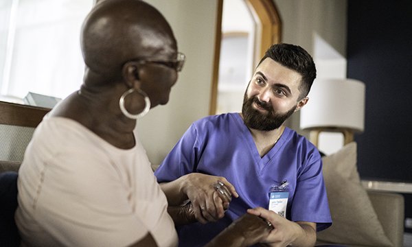 An older woman sitting down holds hands with a nurse about to guide her on standing up