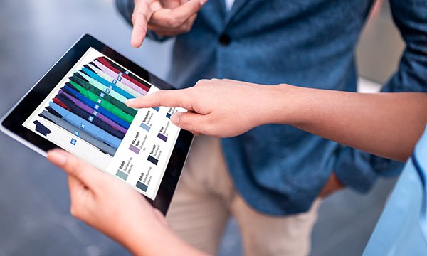 Unseen woman holds tablet computer, man standing alongside. Both pointing to screen displaying image of NHS colour-coding for NHS uniforms
