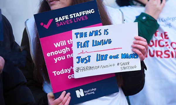 A close-up of a hand-made placard held up by a nurse on a picket line saying 'The NHS is flatlining just like our wages'