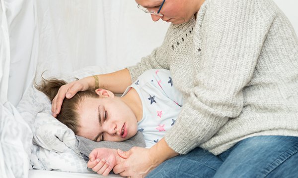 A teenage girl lies on her side looking distressed while her mother sits close by holding her hand and puts her other hand on her head to comfort her