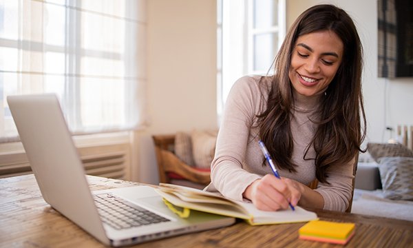Woman smiles as she makes notes in front of her laptop while working at home – doctoral research is a time commitment and work-life balance challenge