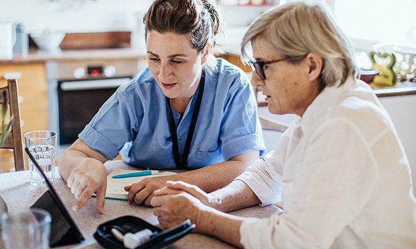 A healthcare professional and older patient run through a blood monitoring process with the aid of a tablet computer at the patient’s home