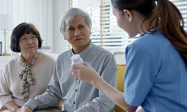 A nurse holds up and shows a bottle of medication to an older man seated next to his female companion