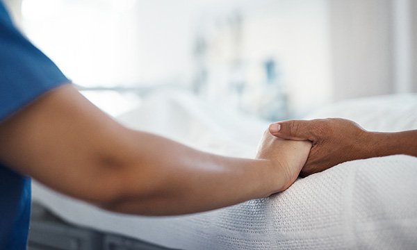 Close-up image of two hands, as a nurse in uniform sits at the bedside of a dying patient in hospital, holding their hand