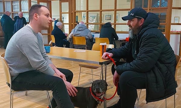 Nursing student Andrew Poole sits at a table in a community hall talking to a parish nursing client while on clinical placement