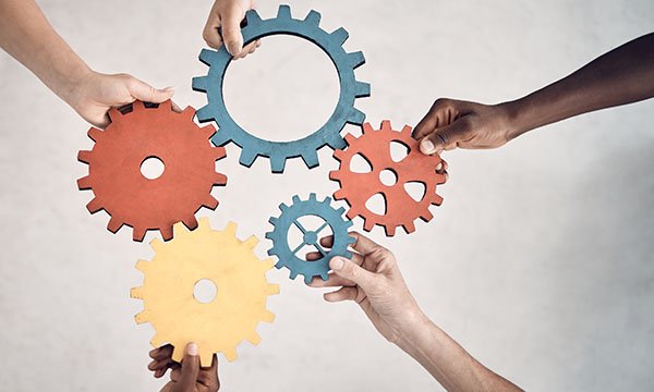 Five arms out-stretched from all sides into centre of the frame. Each hand holds a cog that fit together to illustrate teamwork in nursing