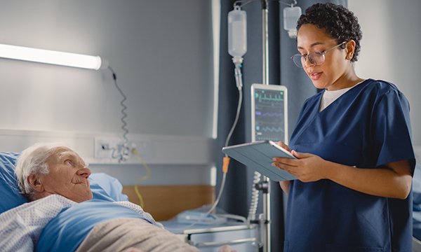 An older man lies in a hospital bed while a specialist nurse records data and assesses his condition