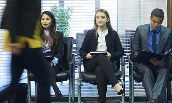 A row of people in smart attire seated in a waiting area looking slightly anxious and preoccupied, as if waiting to be called into a job interview
