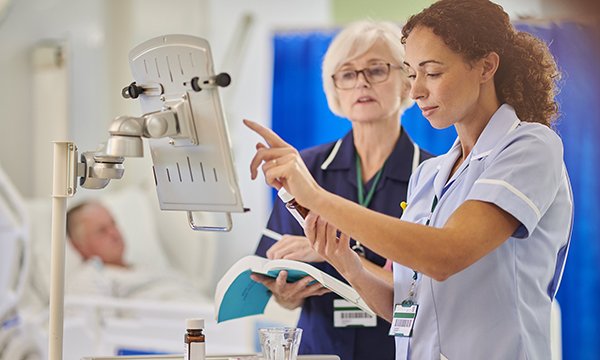 Two nurses, one holding a reference book and the other a bottle of medicine, consult a display board in a ward setting