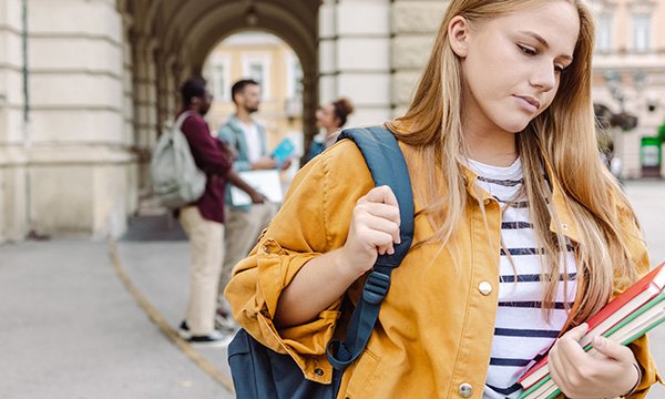 A female student looks downcast and worried as she walks alone outside of a university building while other students chat in the background, suggesting a nursing student who is isolated or struggling with their mental well-being