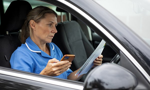 A community nurse sits in her car using her phone while looking through patient notes