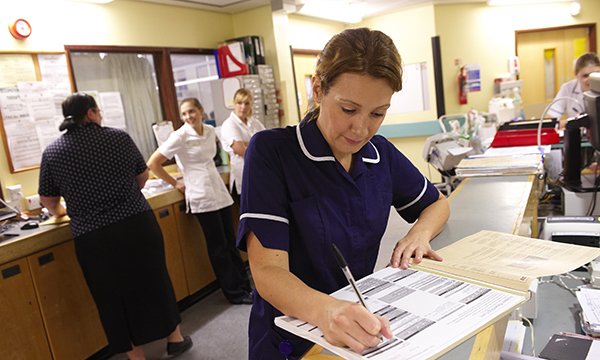 A ward manager fills in a form while at a nurses’ station. Three colleagues are standing in the background.