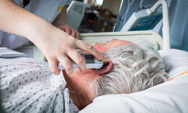 Image of an older male patient lying in a hospital bed having his temperature taken by a nurse using a digital thermometer
