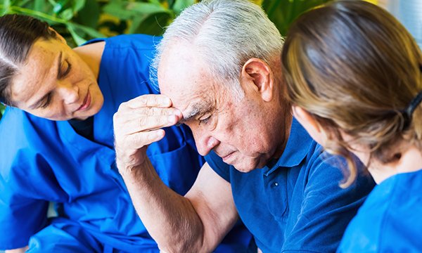 An older man with his head bowed appears pensive as he holds a hand to his forehead, while nurses sitting either side of him watch closely and try to engage him in conversation 