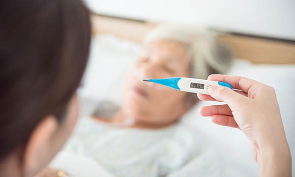Treating high temperature with paracetamol: a nurse with a thermometer showing 39.5°C, after taking the temperature of an older patient lying in a hospital bed