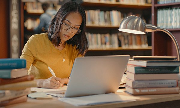 Studying for a master’s degree: a young woman makes notes in a library with a stack of books on her desk