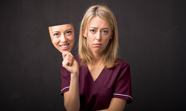 People pleasing can take a toll on your well-being: a nurse holds a smiling mask of her face away from her real face, which looks sad  